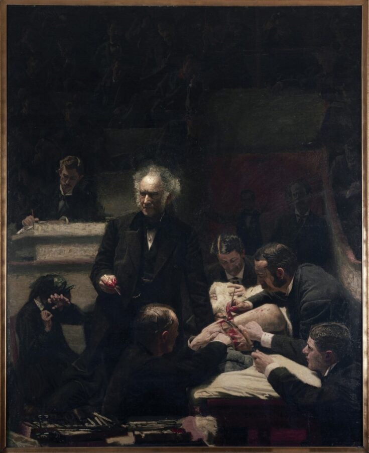 A dimly lit operating theater shows surgeon Samuel Gross standing at center as he oversees a procedure, surrounded by assistants working on a patient. Figures watch from the gallery behind him.