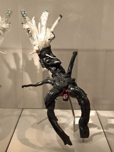 Sculpture from a branch showing a figure with a large silver crown and outstretched arms leaning back to give birth, with the child beginning to emerge.