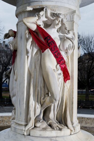 A classical marble statue on a public monument draped with a bright red sash reading “RESIST!"