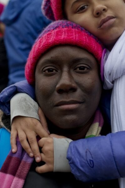 A person wearing a bright pink knit hat looks directly at the camera while a child in matching colors rests against their head, wrapping their arms gently around the adult’s shoulders.