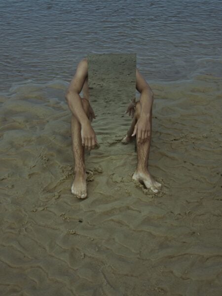 A person sits on wet sand with the ocean behind them, holding a rectangular mirror that hides their torso and head while their arms and legs frame the mirror’s reflection of the sand.
