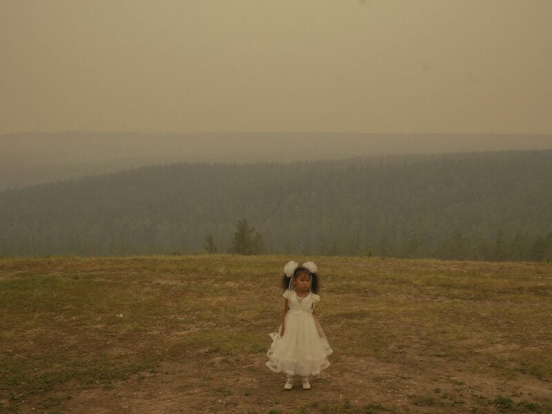 A small child stands alone on a vast prairie in a white dress with matching hair bows, with a hazy view of mountains in the distance.