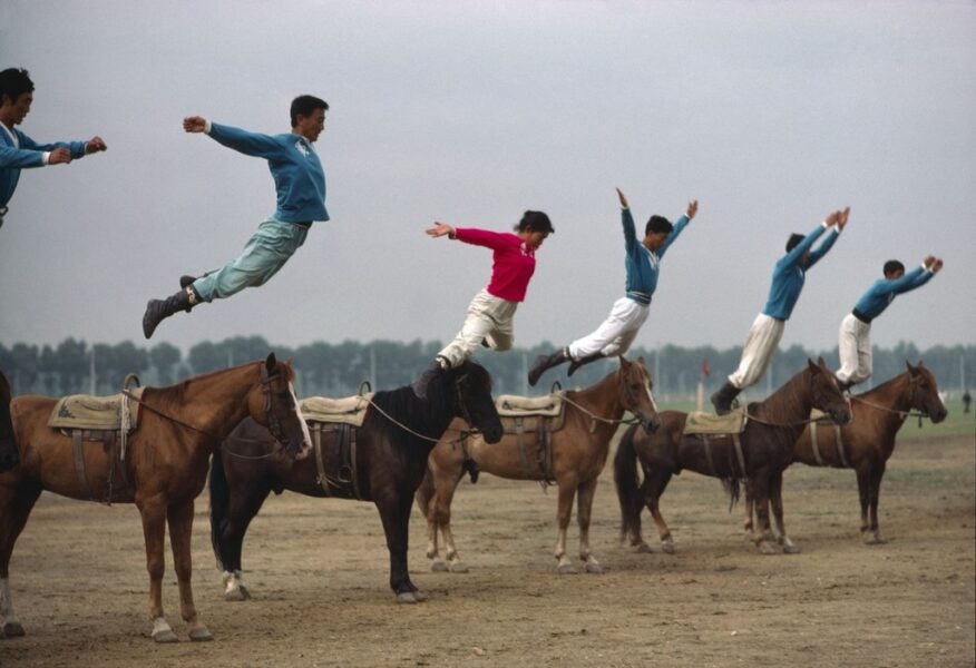 Acrobatic riders leap in unison from the backs of standing horses on an open field, their bodies extended in midair while the horses remain aligned and still.