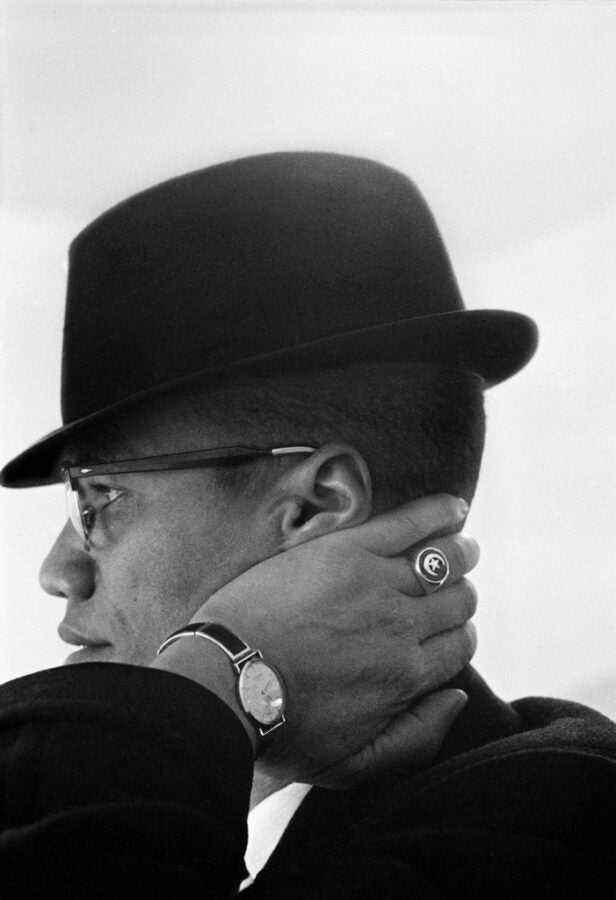 Black and white profile portrait of a man in a brimmed hat and glasses, his hand resting on his neck, wearing a watch and a ring with an emblem.