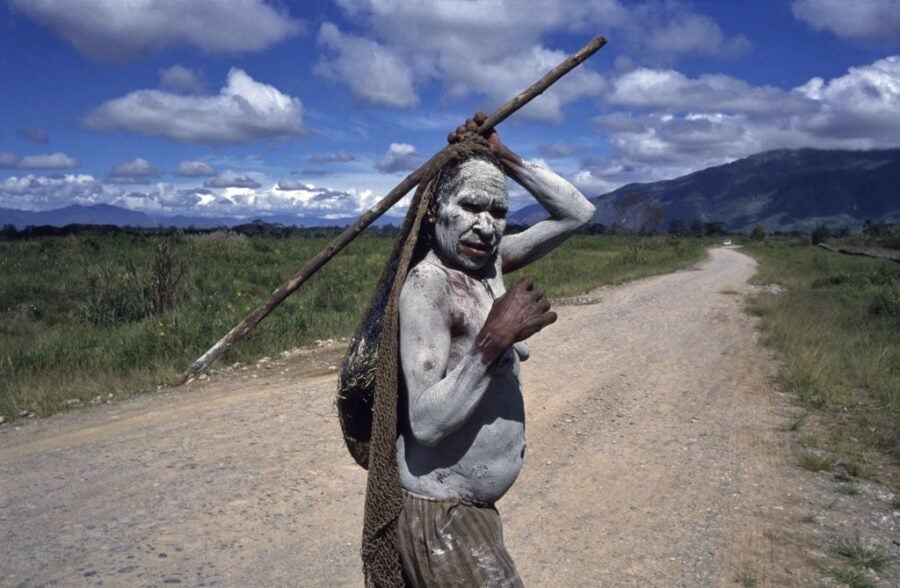 A person covered in white pigment walks along a dirt road in a wide valley with mountains rising in the distance, carrying a long wooden pole.