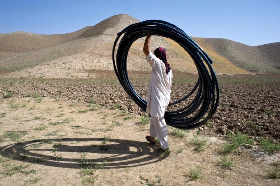 A person in loose-fitting clothing with a red head covering walking away from the camera into a desert landscape, carrying a large bundle of black plastic hose.