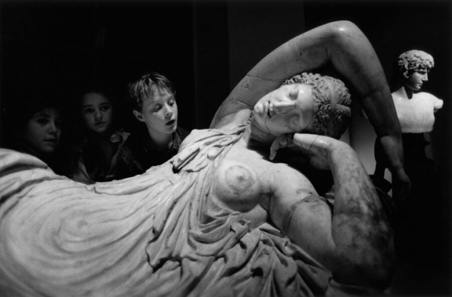 Three children view a large classical marble statue of a reclining woman in a loosely draped chiton, her eyes closed and one arm resting over her head, while one child leans in to study the torso closely.