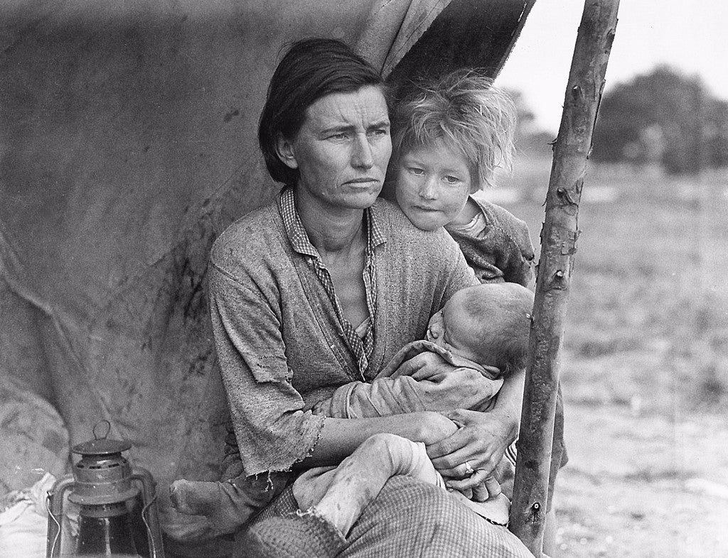 Black-and-white photo of a woman with a careworn expression cradling an infant in her lap under a makeshift tent. A child with wild, uncombed hair leans over her shoulder. All are dirty and wearing ragged clothing.