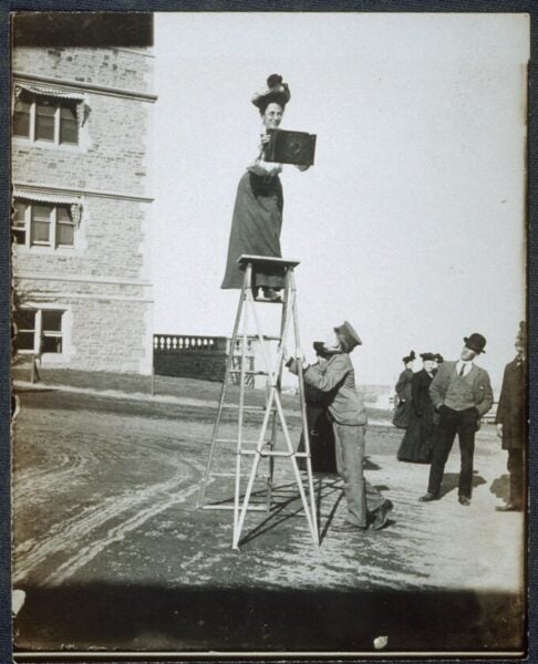 A woman in early 1900s dress balances on the top step of an eight-foot ladder and uses both hands to frame a picture with a large camera. Below her, a man balances the ladder and spectators look on.
