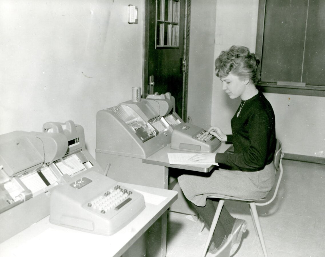A black and white photo of a women seated at an old and large computer, typing during the mid-20th century.