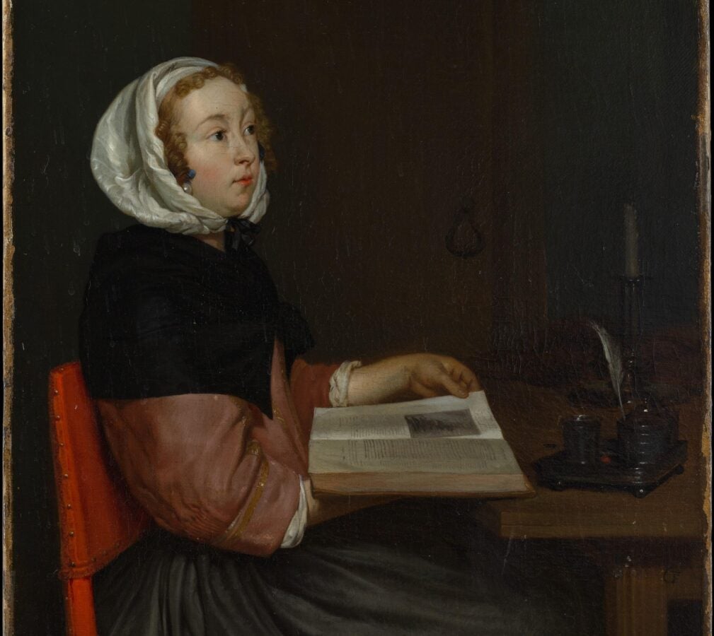A painting of a woman from the seventeenth century. She sits at a desk reading a book.
