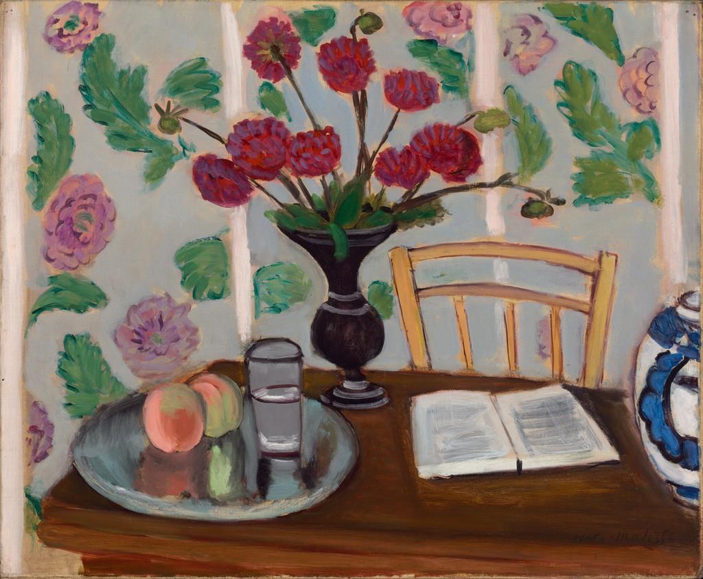 This is a vibrant still life painting by Henri Matisse from 1923. It depicts a wooden table set against a patterned wallpaper of green leaves and pink flowers. On the table, there's a dark vase with bright red blossoms, a plate with a peach and a glass of water, and an open book beside a blue and white decorative pot. A wooden chair is placed against the table, inviting viewers into this cozy, domestic scene.