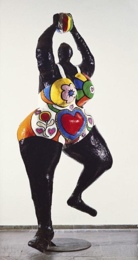 Large black sculpted figure dancing on one leg, wearing a brightly painted bodice with hearts, flowers, and bold colors, holding a patterned sphere overhead.