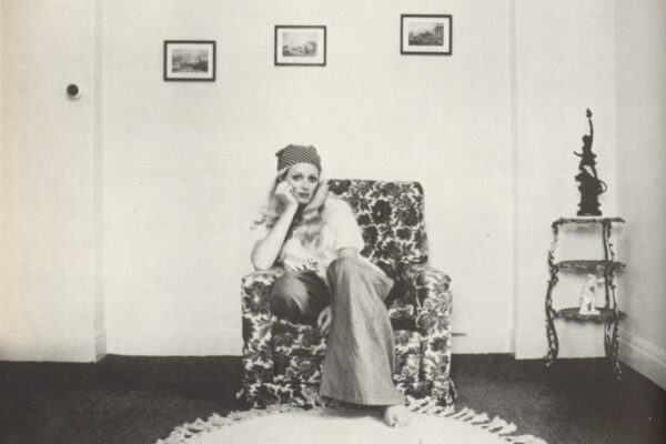 A black-and-white photograph of a woman posed seated in a chair against an wall of an apartment.