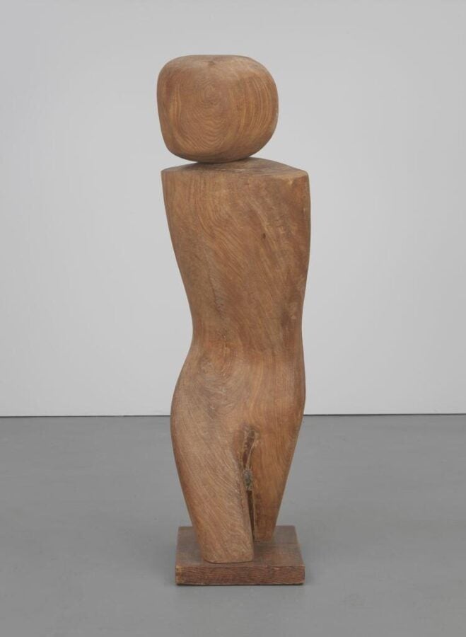 A smooth wooden sculpture resembling an abstract human figure with minimal curves, standing on a simple wooden base against a gray wall.