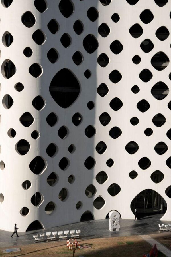 A modern white building facade with a pattern of rounded diamond-shaped windows. Small figures of people walk near the base, emphasizing the scale of the structure.