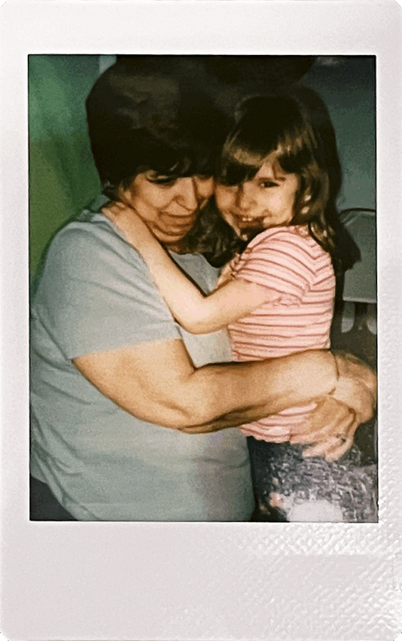 A woman with short dark hair hugs a young girl in a striped shirt. The girl smiles shyly while being held close in a warm, affectionate embrace.