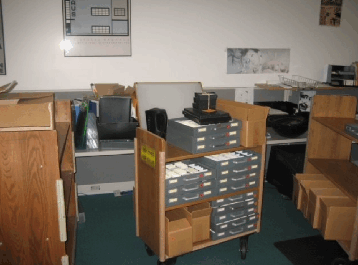 A wooden cart holding trays of photographic slides, surrounded by office furniture and boxes in a workspace.