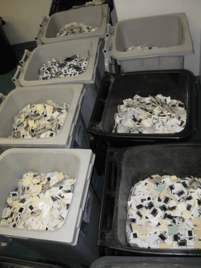 Large bins filled with thousands of discarded photographic slides and paper slide mounts arranged in rows in a storage room.