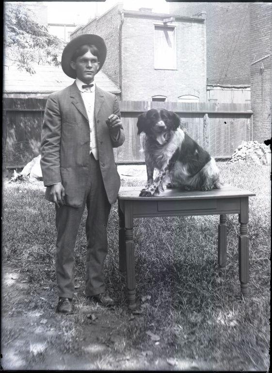 A man in an old-fashioned suit and hat stands beside a dog sitting on a table in a grassy yard.