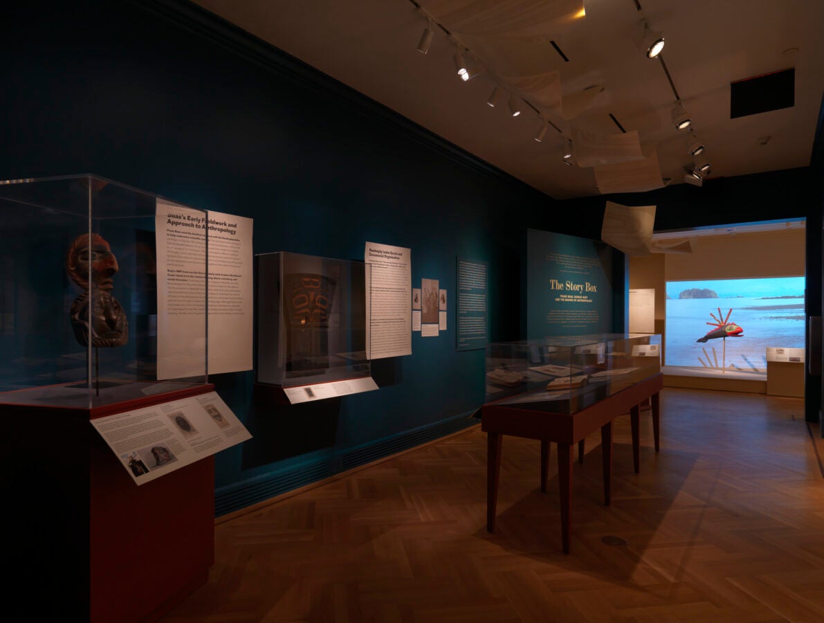 A dimly lit museum gallery with teal-blue walls displaying Indigenous art and artifacts in glass cases. Text on the wall reads “The Story Box,” and one display case holds carved masks. Through a window at the back, a coastal landscape with water and small islands is visible.