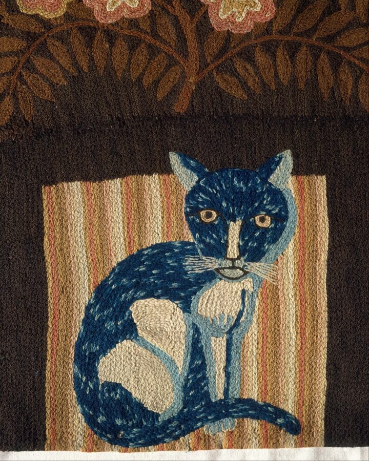Folk-art embroidered carpet panel featuring a blue, wide-eyed cat sitting on a striped ground.