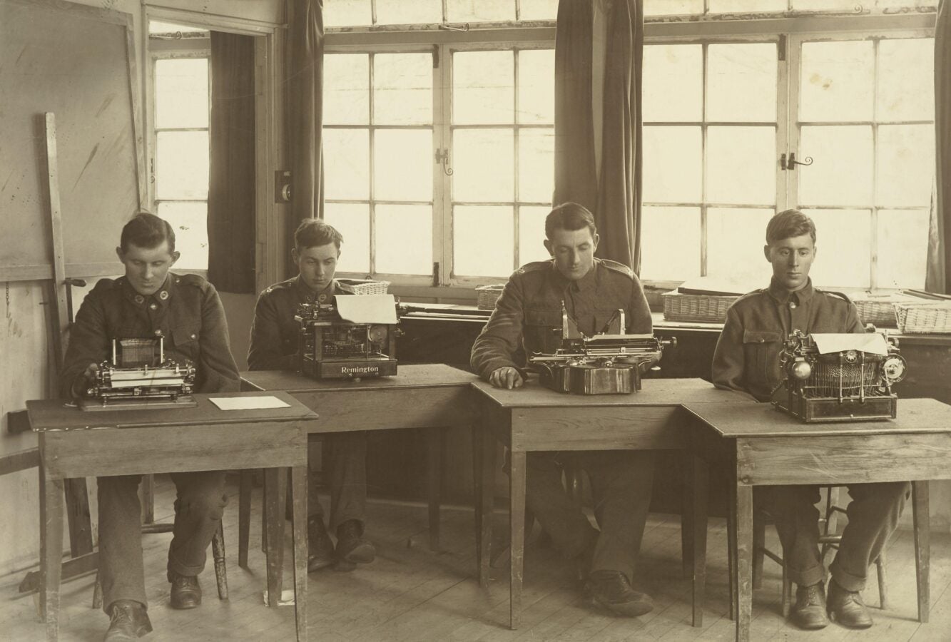 A black-and-white photograph of four young men seated at separate wooden desks, each typing on a typewriter in a sunlit room with large-paned windows behind them.