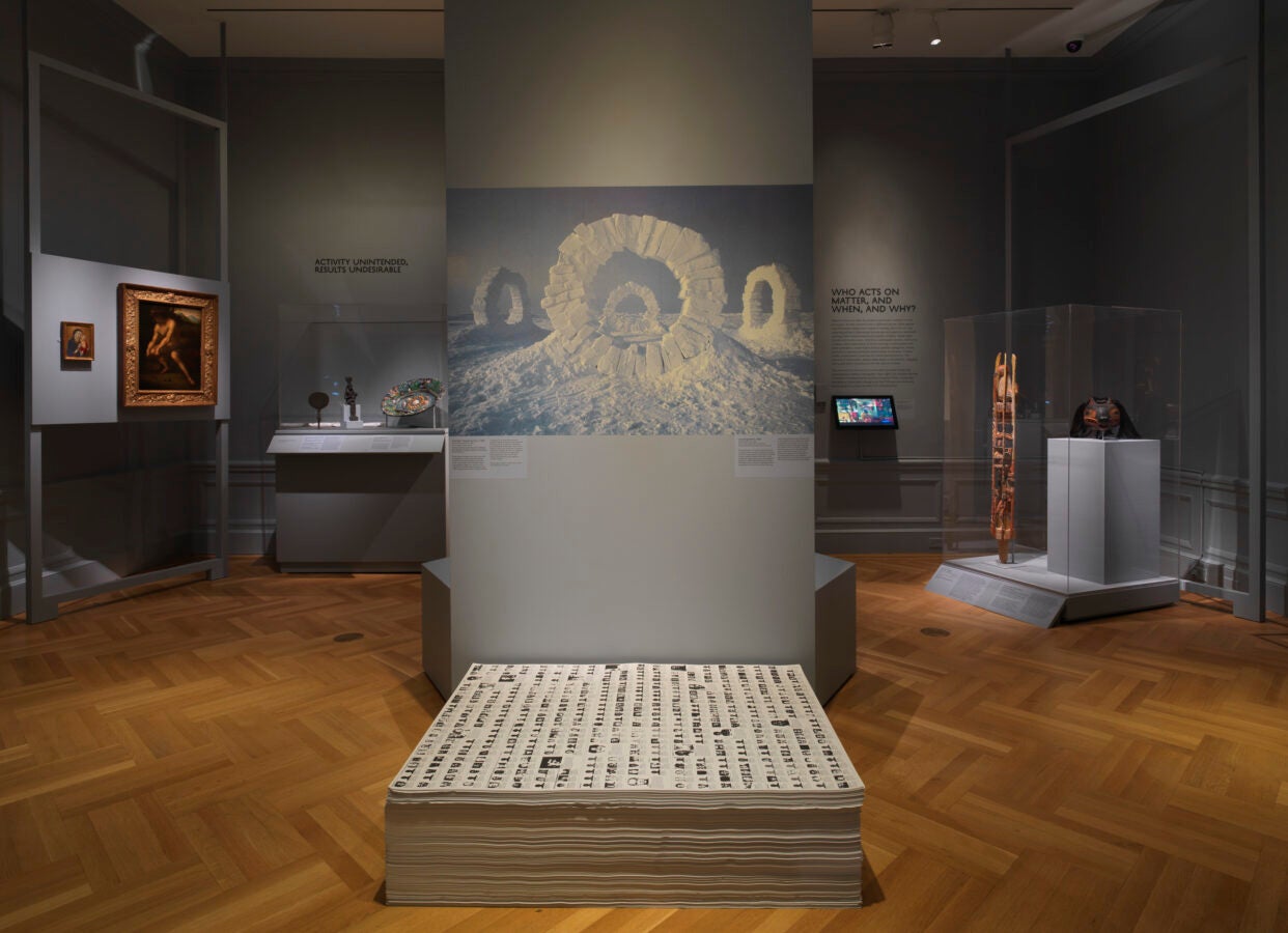 A contemporary museum gallery with gray walls showcasing diverse artworks, including a large photograph of snow arches, a stack of printed sheets in the foreground, and various sculptures and paintings displayed in glass cases and on walls around the room.