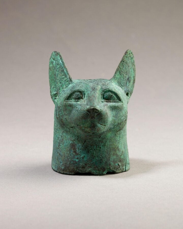 Ancient bronze cat head with upright ears and almond eyes, set against a neutral background.