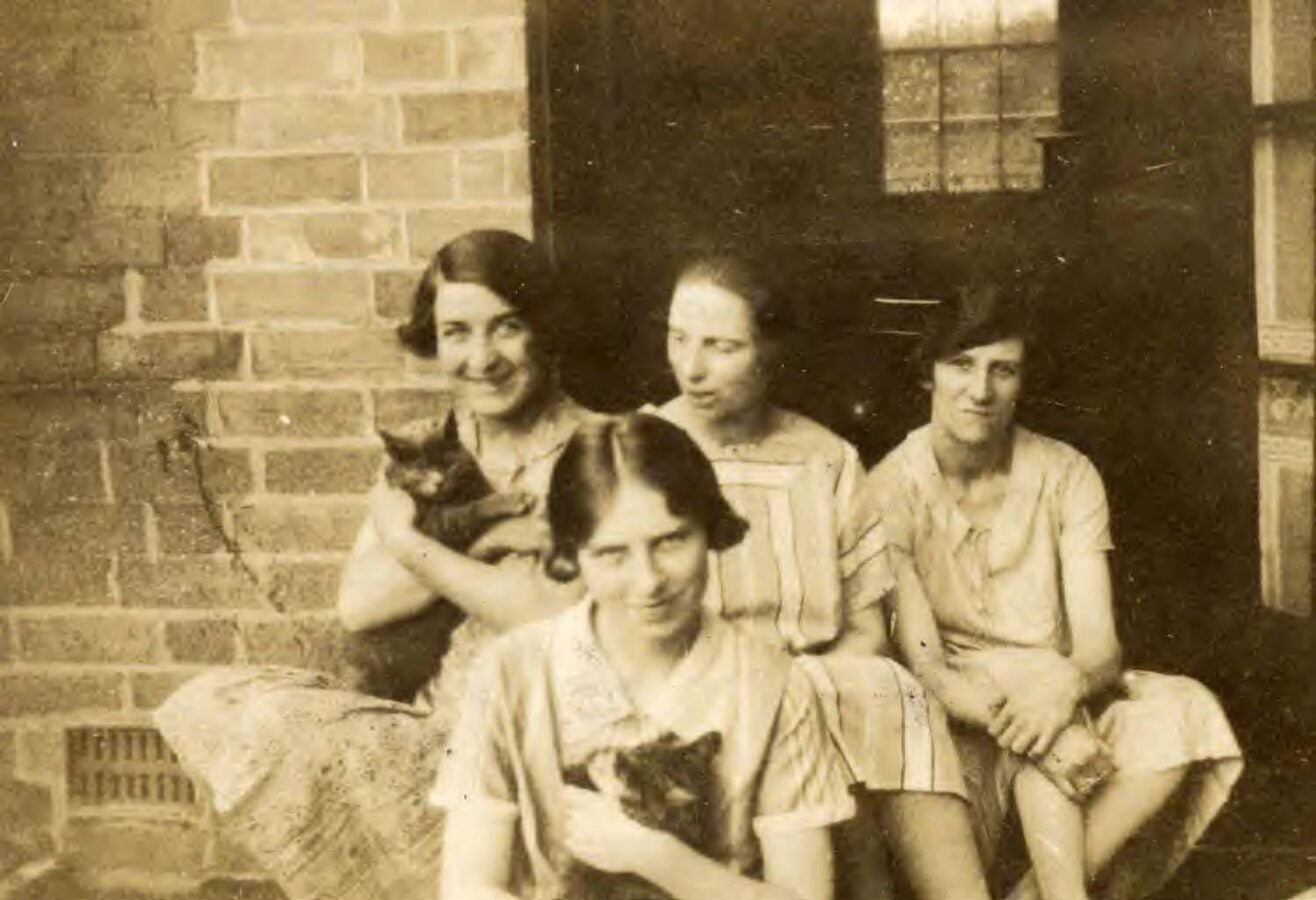 Vintage photo of three women seated on a porch, each trying to hold a squirming cat for the camera.