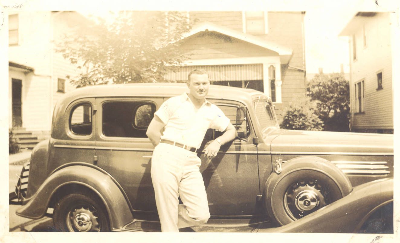 Baseball player Johnny Mize leans casually against a vintage car in a residential neighborhood, wearing a short-sleeved shirt and slacks.