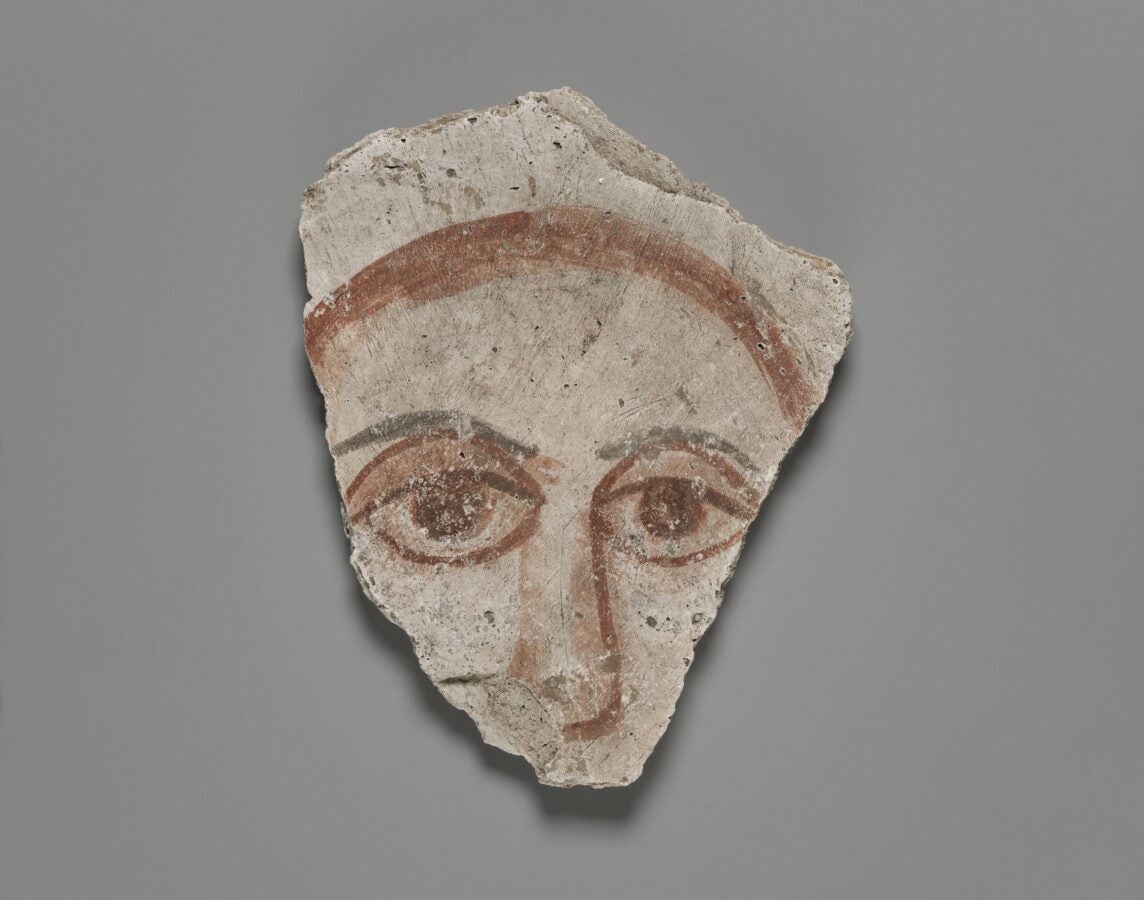 Painted plaster fragment with a stylized face: wide almond-shaped eyes and a curved reddish brow line on a gray background.