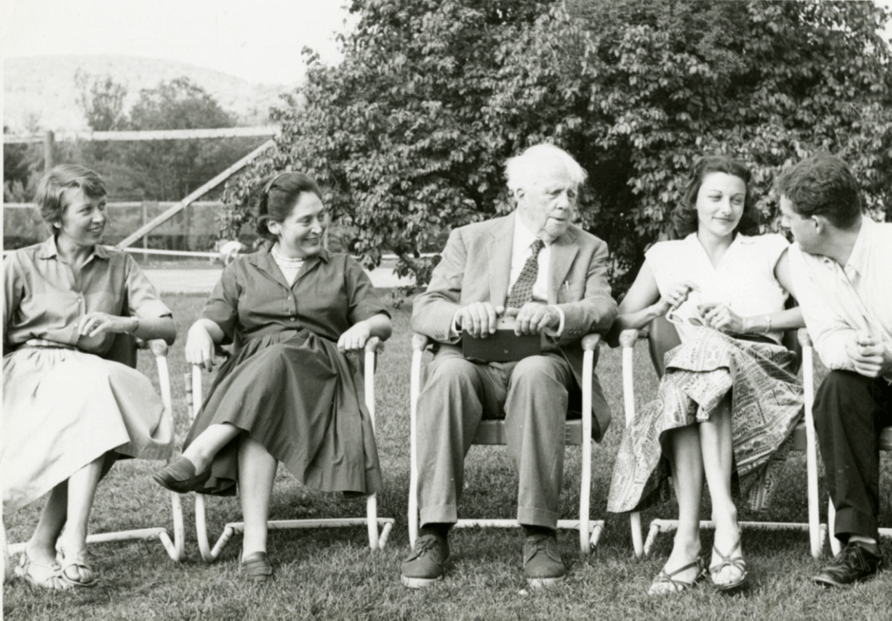 Poet Robert Frost sits outdoors in conversation with Anne Sexton and other attendees at the Bread Loaf Writers’ Conference, surrounded by chairs and trees.