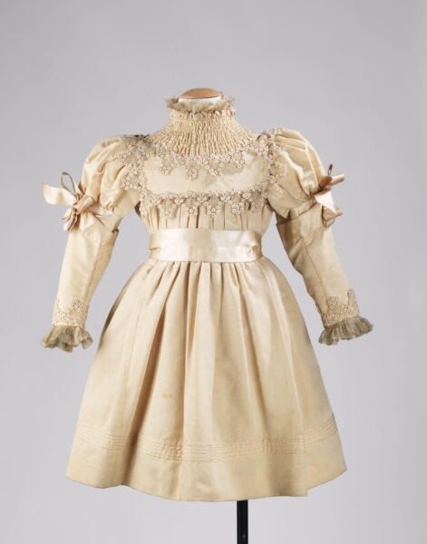 A vintage 1895 girl's party dress with an ivory hue. The dress features intricate lacework around the neckline, sleeves, and bodice, with a smocked high collar. Puff sleeves are adorned with ribbon bows, and the skirt has subtle pleats and embroidery near the hemline.