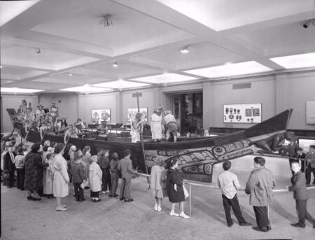 A black-and-white photograph depicting a group of children and adults gathered around North West Coast Canoe displayed in a museum setting. The canoe features detailed artwork with traditional motifs, and several lifelike mannequins dressed in ceremonial attire are positioned in and around the canoe, recreating a historical or cultural scene. The museum walls display additional artifacts and exhibits, creating an educational environment.