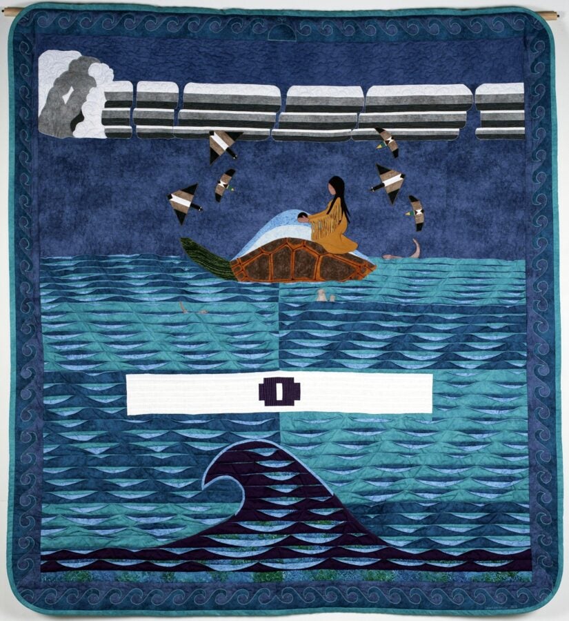 Textile artwork depicting a woman sitting on a turtle in the ocean beneath a blue sky with broken white beams above.