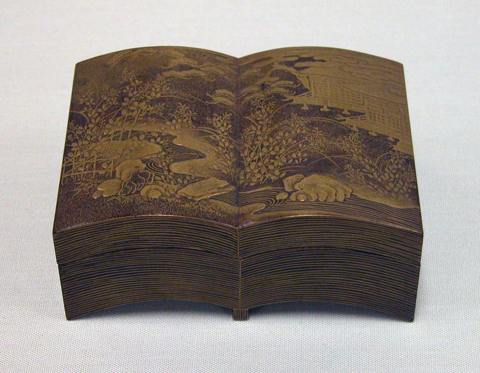 A decorative Japanese box from the late 19th century, shaped like an open book. The box features intricate lacquer work depicting a serene landscape, including a pond with rocks, delicate plants, and a traditional structure in the background. The craftsmanship captures fine details, giving the impression of a textured scene etched onto the box’s surface.