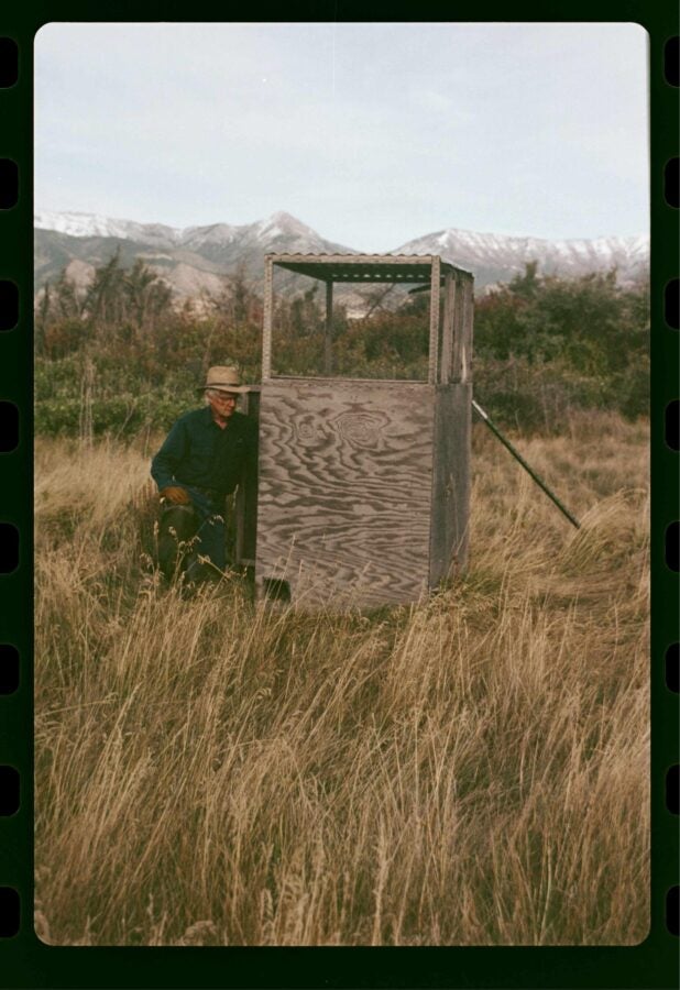 A man kneels beside a small wooden observation hut in a grassy field with snow-capped mountains in the background.