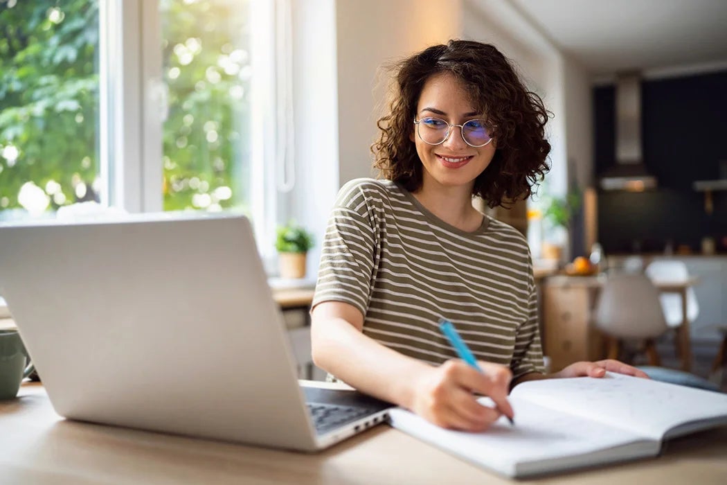 A young woman with curly hair and glasses is smiling as she writes in a notebook while sitting at a desk with her laptop open. She is in a bright, cozy room with a view of trees outside the window, creating a comfortable study environment.