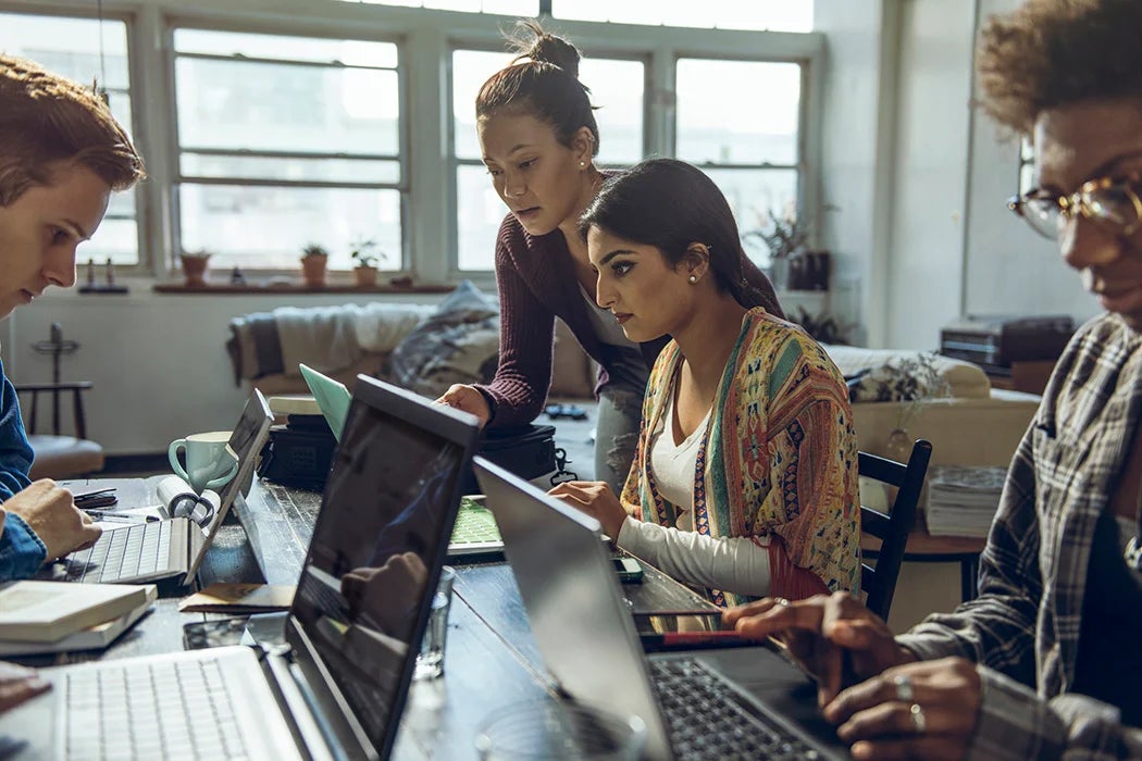 A diverse group of students sitting around a table, focused on their laptops and notebooks. They appear to be collaborating on a project in a sunlit room, fostering a setting of teamwork and shared learning.