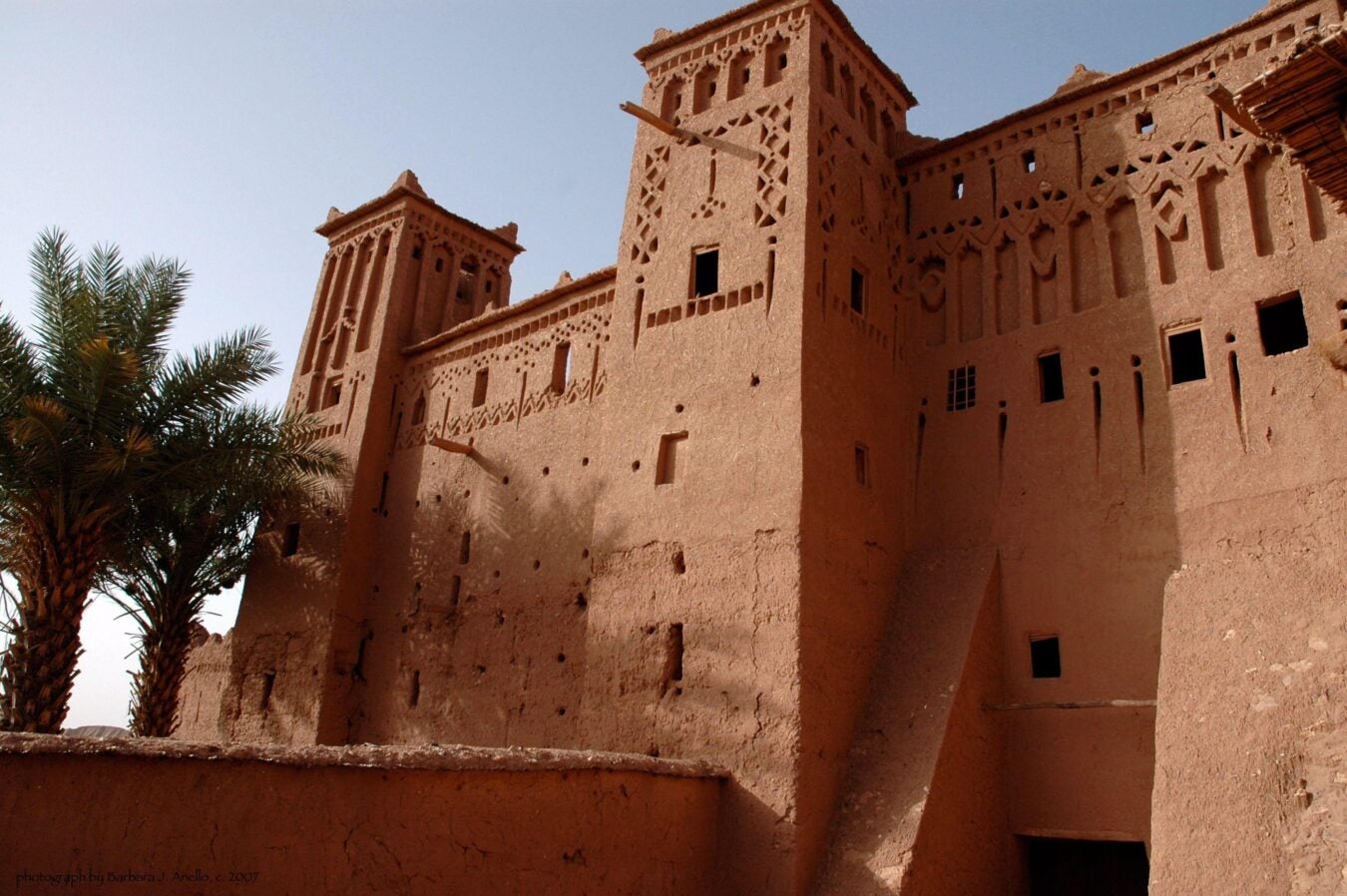 View of the Ait Ben Haddou kasbah, a traditional Moroccan earthen clay architecture structure with high towers and intricate geometric patterns. The warm-toned walls contrast with the bright blue sky, while palm trees add a natural element to the scene. The structure's defensive design and decorative details highlight its historical and cultural significance.