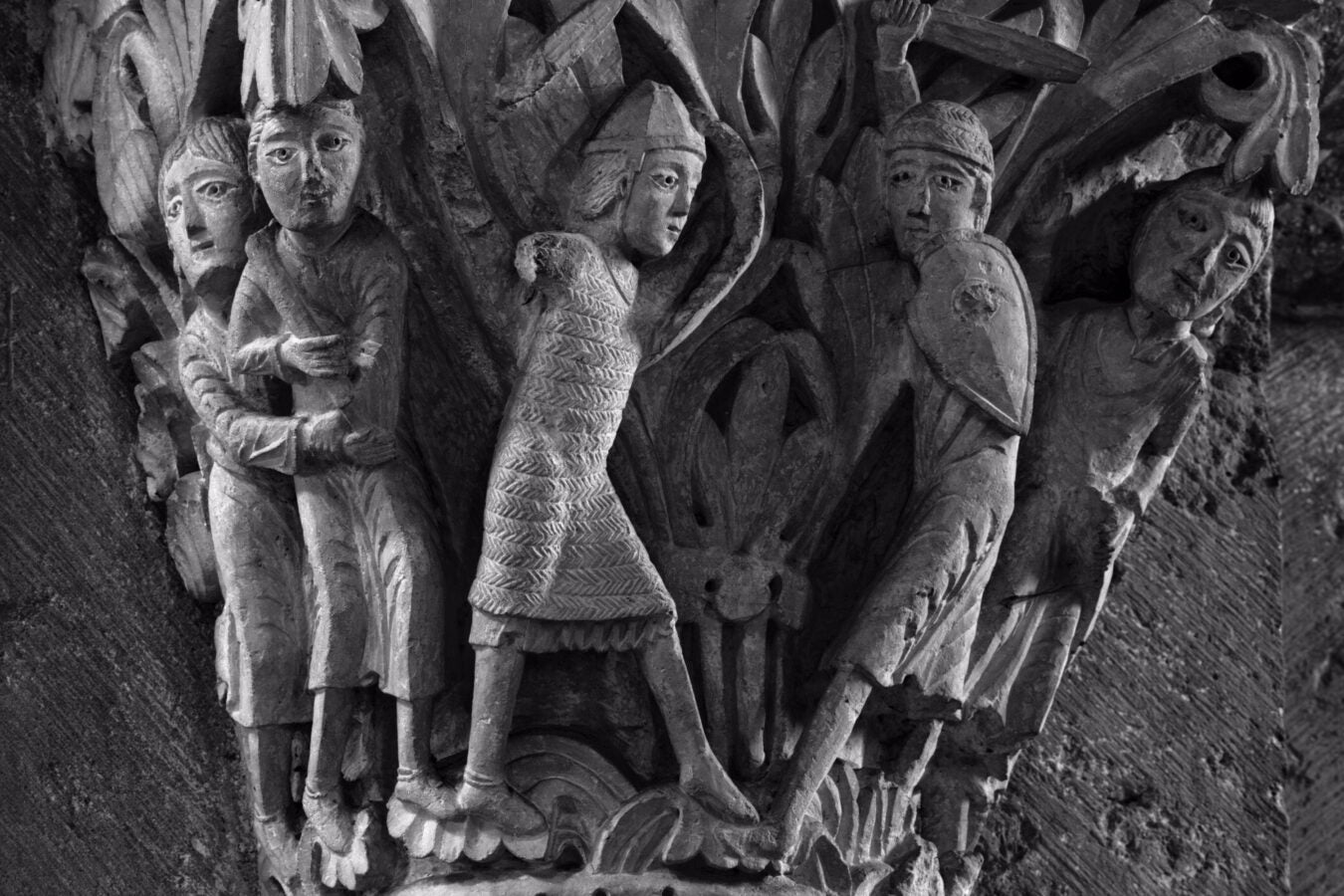 Close-up of an intricately carved capital from the Basilique Sainte Madeleine, depicting detailed figures in a narrative scene. The figures include soldiers, a central robed individual, and others engaged in expressive postures, surrounded by stylized foliage and architectural elements. The carving showcases the craftsmanship and storytelling typical of early Romanesque art from the 11th century.