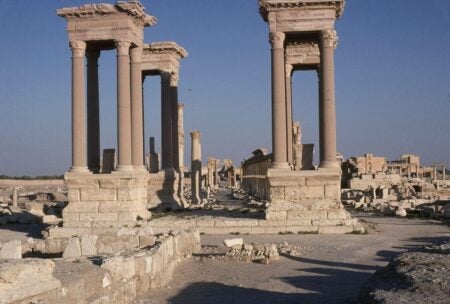 View of the Tetrapylon in Palmyra, Syria, an ancient Roman monument featuring four groups of tall columns topped with ornate capitals and lintels. The structure stands amid a vast expanse of ruins, with additional colonnades and remnants of buildings visible in the background. The scene is illuminated by natural light under a clear blue sky, emphasizing the grandeur of the archaeological site.