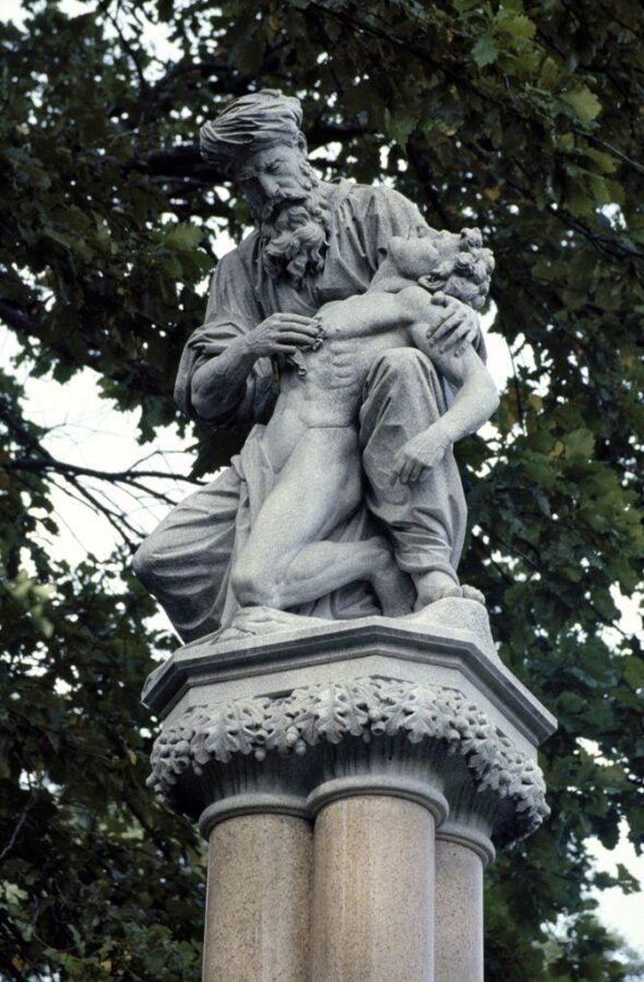 A detailed photograph of a historical statue in Public Gardens near Commonwealth Avenue entrance, Boston, MA, depicting a compassionate figure, likely a healer or doctor, cradling an unconscious or suffering individual. The statue is mounted on a column adorned with carved oak leaves, symbolizing strength and endurance. The figures are sculpted with intricate detail, emphasizing emotion and care. The background features lush green trees, adding a natural setting to this commemorative artwork, likely honoring medical advancements or humanitarian efforts.
