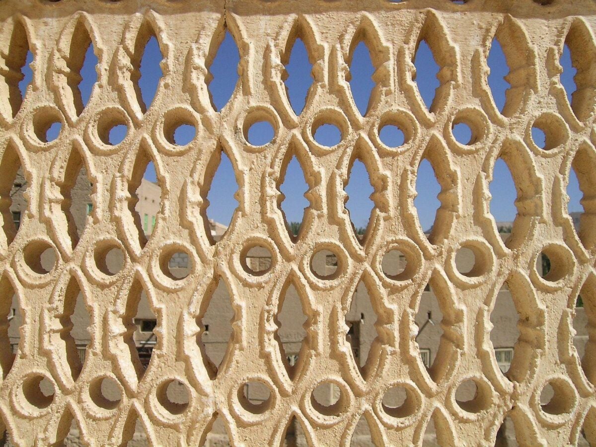 Close-up of Alawi Abu Bakr al-Kaf, an intricately carved lattice screen, featuring geometric and organic patterns with oval and pointed openings. The cream-colored plasterwork highlights the craftsmanship and traditional design, with a clear blue sky and blurred architectural structures visible through the openings in the background.