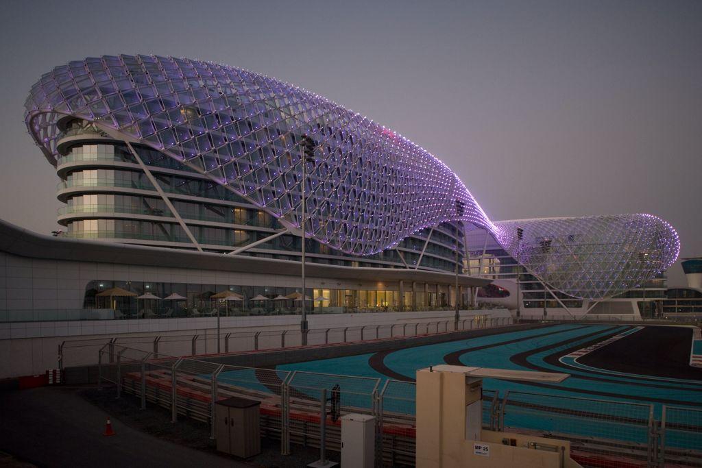 The Yas Hotel in Abu Dhabi at dusk, featuring its futuristic design with a grid-like LED-lit canopy that wraps around the structure. The adjacent Yas Marina Circuit racetrack is visible in the foreground, with vibrant blue and red markings.