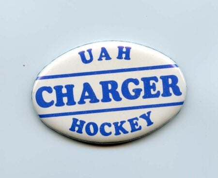 This is a circular button featuring the text "UAH Charger Hockey" in bold blue lettering, with two horizontal blue lines separating the words. The design is set against a white background, likely representing the University of Alabama in Huntsville's hockey team. The button's vintage style suggests it is a memorabilia item, possibly from the 1980s.