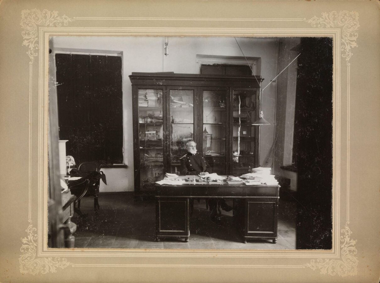 A black-and-white photograph of I.P. Pavlov seated at his desk in the Imperial Military Medical Academy, St. Petersburg, 1904. Pavlov is dressed in formal attire and surrounded by stacks of papers. Behind him, there is a large cabinet displaying various scientific instruments and books. The room has a minimalist design with high windows and a hanging light fixture. The photograph is framed with ornate detailing around the edges.