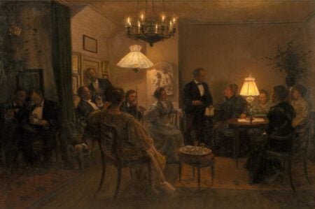 An oil painting by Viggo Johansen titled An Evening Party in the Artist’s Home (1899). The artwork depicts an intimate indoor gathering with elegantly dressed men and women in a warmly lit room. Guests are seated in small groups, engaged in conversation and listening to a man standing while speaking. The room is illuminated by chandeliers and table lamps, creating a cozy and inviting atmosphere. The scene reflects a sense of refinement and social interaction typical of the time period.