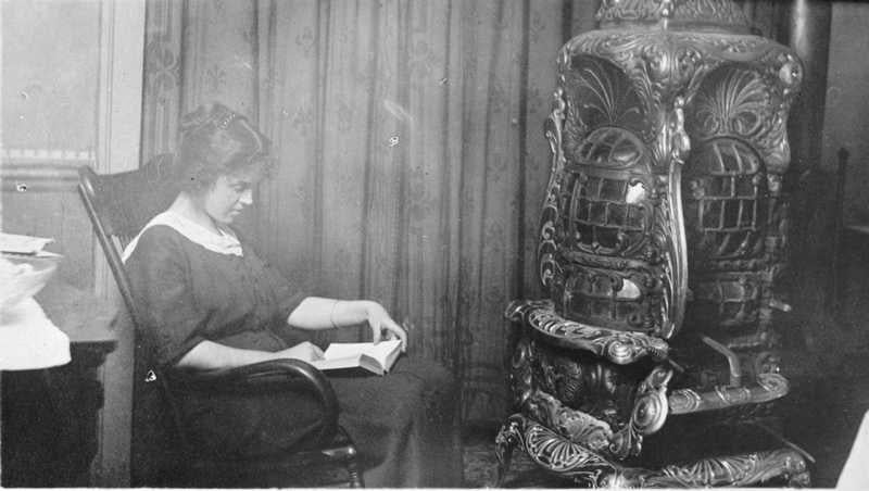 A black-and-white photograph of a woman seated in a wooden chair, reading a book next to an ornate cast-iron wood stove. She wears a dark dress with a white collar, and her hair is styled in a short, wavy bob. The background features a curtain and part of a table with a white cloth. The stove has intricate detailing with decorative patterns and a polished surface. The image conveys a quiet, intimate moment of reading and warmth.
